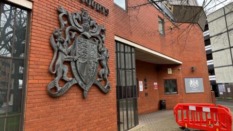 The outside of Swindon Crown Court. It is a modern red-brick building, with large windows and a very large metal coat of arms on the wall. 