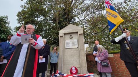 A war memorial in Shrub End near Colchester is rebuilt and unveiled. The Bishop of Chelmsford reads a short sermon. Standing next to the memorial is Sir Bob Russell, High Steward of Colchester, and Sue Starling, widow of Kevin Starling who campaigned to have the memorial re-built before his death in January 2024