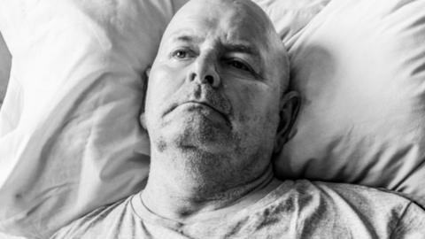 Black and white image of John Newcombe, lying in bed with his hand on his chest, starring into the distance.