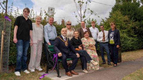 Wolverhampton mayor Craig Collinswood is sitting on the memorial bench. He is wearing his chain of office and a dark suit. He is surrounded by a group of people who are the friends and family of former councillor Paddy Bradley