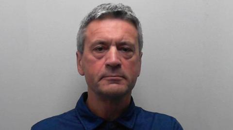 A man faces the camera against a plain light-grey background. He has short grey hair and is wearing a dark blue collared shirt.