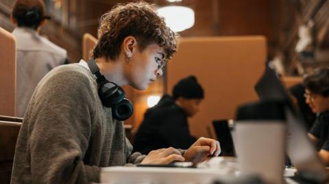 A person with short curly hair types on a laptop at a wooden desk in a library and study space, wearing glasses and headphones, with other people working nearby in the background.
