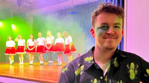 Jordan Winn stands in the foreground with a line of dancers in identical white tops and red skirts in the background. Jordan is wearing green make up on his eyes and a black shirt.