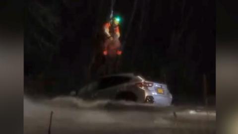 Two people are airlifted from above a car half-submerged in water at night.