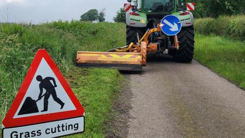 A red road sign reads 'grass cutting' and has a person digging into the ground. The sign is place behind a tractor which has a grass cutting strimmer fixed to its left side. It is a small single-track road which has had its verge partly cut. 
