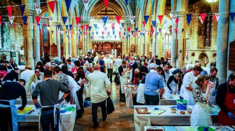 Lots of people in aprons leaning over tables in a church judging pies