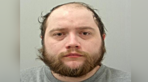 Custody photo of Dylan Saville wearing a grey sweatshirt while against a light grey background. He has lots of brown facial hair and brown eyes. 