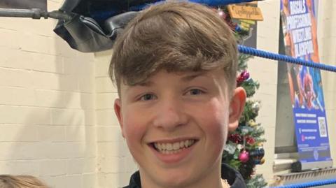 Charlie has brown hair and is in a corridor with white painted bricks.