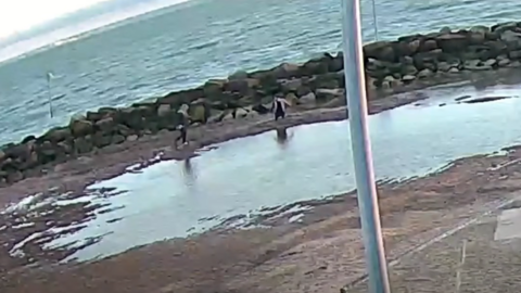 A lagoon with two people standing next to a pile of mud. There is water slightly further afield and a silver pole also in the camera shot.