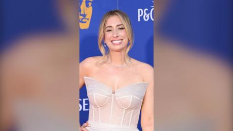 A woman, Stacey Solomon, wearing a white dress against a blue background, smiling to camera.