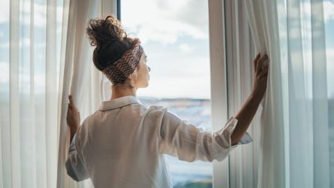 A young woman closes the curtains by a window.