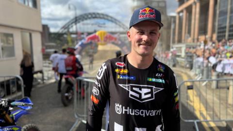 Head and shoulder shot of a smiling young man with a moustache. He is wearing a much-branded long-sleeved t shirt and Red Bull baseball cap. Behind him, and slightly out of focus, can be seen the Tyne Bridge, with a race entrance arch, and crowds lining the street.