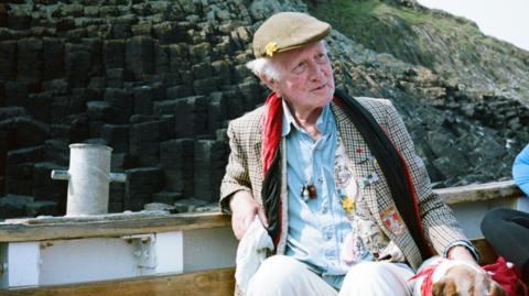 Peter Monro. He is an elderly man sitting on a boat in front of some cliffs. He is wearing a flat cap, a blue shirt and a plaid blazer featuring various patches of colorful fabric, and a red velvet scarf. He is looking to the side away from the camera.