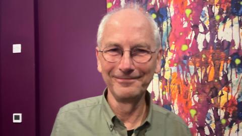 A man with receeding grey hair and glasses smiles. He is wearing a moss green shirt and standing in front of a brightly-coloured abstract painting hung on a purple wall.