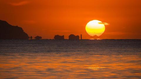 A vibrant orange sky surrounds a bright yellow Sun as it rises over the sea, headland and rock stacks
