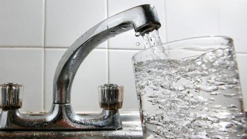 A glass of water is being filled from a silver tap. Water can be seen coming out of the spout and filling up a clear glass.