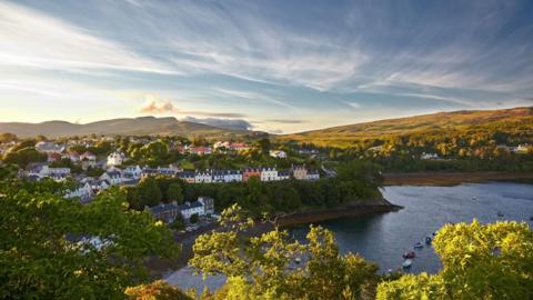 Port Rìgh. Baile an cois a' chladaich le craobhan agus beanntan fada às. 