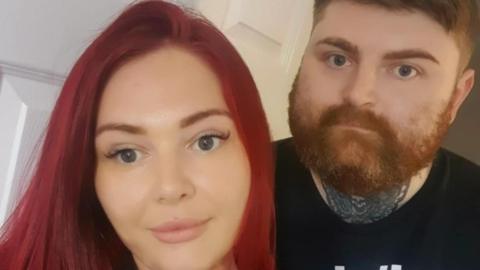 Emily Luccarini smiles at the camera. She has long dyed red hair. Her brother stands behind her smiling. He has a thick reddish beard, neck tattoo and he is wearing a black T-shirt.