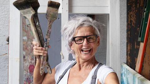 A grey haired woman wearing glasses in a white t-shirt and grey dungaree, holding 3 paintbrushes in one hand and two in the other. 