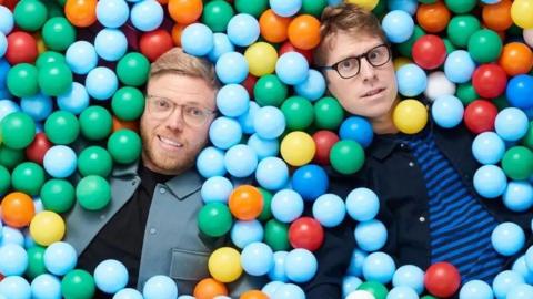 Rob and Josh lie in a colourful ball pit, with their bodies mostly submerged. Their faces peer out and they are looking directly at the camera.