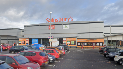 The outside of a large Sainbury's supermarket entrance with a full car park