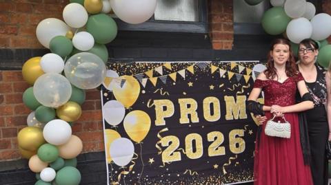 Lillie-Mae is standing to the right of the picture in front of a black sign with the words 'Prom 2026' in gold lettering. The sign also has white and gold balloons and bunting on it. Lillie-Mae is wearing a red off the shoulder dress with a black shawl draped over both arms. She is carrying a white handbag. To her left is her mum Katie, who is wearing a black top with a multi-coloured pattern.