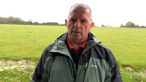 A head and shoulders photo of a man with grey hair wearing a green and black waterproof jacket. There's a large expanse of grassland behind him, with a line of trees on the horizon. The sky is cloudy and his coat is wet with rain.