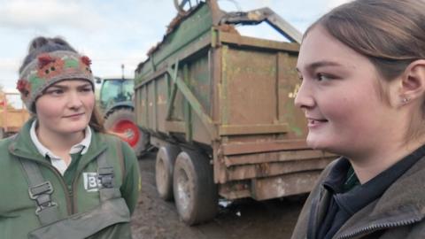 Two women stand talking in the farm yard in front of a tractor and trailer