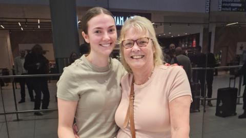 A younger woman with brown hair and wearing a plain khaki t-shirt stands close to an older blonde woman in a pink t-shirt at the arrivals area of Manchester Airport. Both smile and look at the camera.