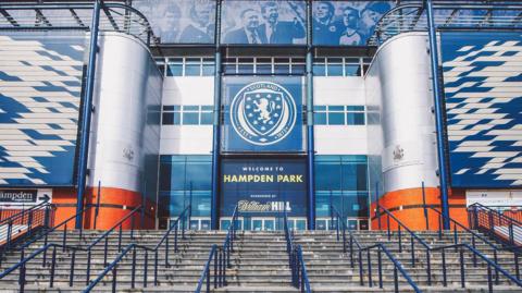 The entrance of Hampden Park, which has grey stairs leading up to the front doors with blue hand rails. The building is grey, blue and orange.