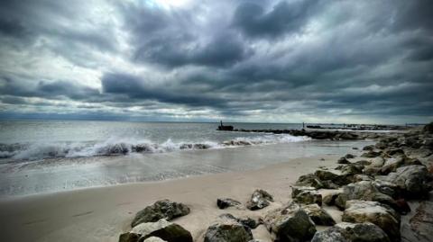 The waves are crashing on to the beach while the sky darkens overhead. On the beach, there are also several boulders.