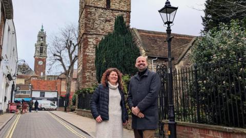 Colchester City Councillor, Natalie Sommers, with Mark Jarman-Howe. They are stood on a narrow road in front of Holy Trinity Church. Ms Sommers has curly hair and wears a beige dress with a rolled neck and black coat. Mr Jarman-Howe has facial hair and wears a long grey coat and brown trousers. 