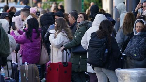 A crowd of people wearing coats and some holding their luggage stand outside the airport.