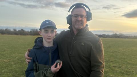 Lucas holds the sword pommel as he stands next to his dad Dave, who is wearing a pair of headphones