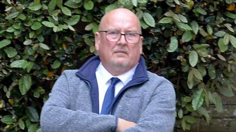 A bald-headed man with glasses and a goatee beard in a blue fleece top with white shirt and blue tie underneath. There is a hedge behind him.