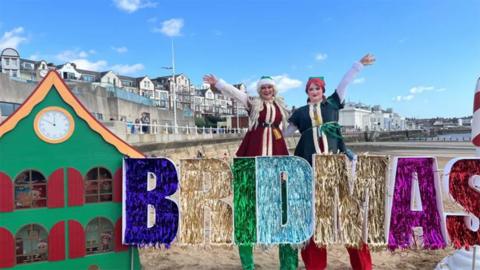 Bridmas day celebration with two stilt walkers dressed as elves on Bridlington beach standing next to a green, wooden house in front of a tinsel Bridmas sign 