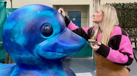 A woman in a brown apron is painting the head of a 1.5m high duck statue that has been decorated in a marketplace entrance. The woman has long blonde hair and a pink and black jumper. The duck is painted in blue and green swirls with stars also painted on. Behind her is a leafy area of the market and a blue door leading to the toilets.