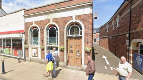The image shows the street‑front exterior of a single storey red brick building. Its façade features three tall, arched window openings. The central bay contains a wooden double door with rectangular panels and metal letterboxes. Hanging flower baskets are mounted beneath the windows. Several pedestrians are visible on the pavement directly in front of the building.