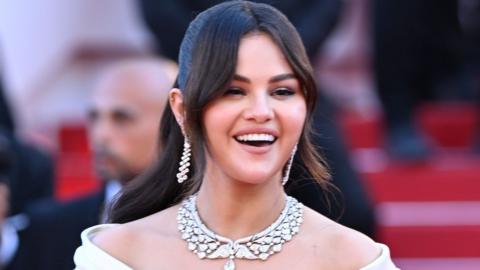 Actress Selena Gomez smiles on the red carpet at the Cannes Film Festival. She wears a white dress with exposed shoulders, with a diamond necklace and earrings. Her long dark hair is held back with a hairband.