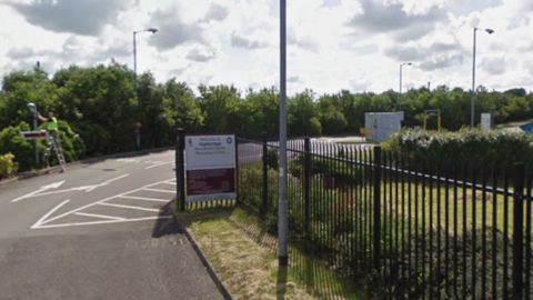 A google image view of the entrance to a recycling centre. There is a sign on green railings and grass verges. In the background is a man wearing high vis on a ladder.