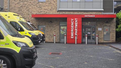 A brown brick building with red wall in front of the door. It says "EMERGENCY DEPARTMENT ENTRANCE" on. There are three ambulances parked in front of it. 