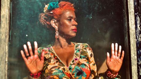 Singer Carmen Souza dressed in a floral dress looking out of a window with an old frame. She has her hands pressed up against the pane. She is wearing red lipstick, has flowers in her hair, long amber-looking earrings and red beaded bracelets on both hands