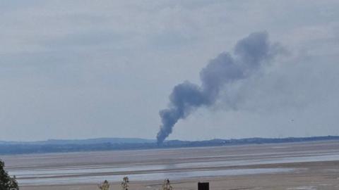 A large plume of smoke can be seen billowing above the sands of Morecambe Bay with hills in the background.