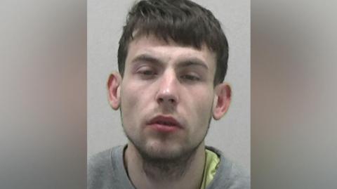 Mugshot of Dallan Tearse. He has thick dark hair, faint stubble and a glazed expression on his face with his eyes half closed. He is wearing a grey sweatshirt