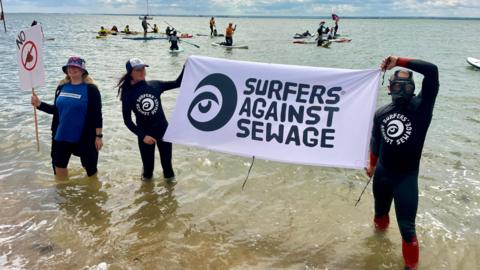 People wearing wetsuits stand in shallow seawater holding a banner reading “Surfers Against Sewage”, while paddleboarders and kayakers gather offshore behind them.