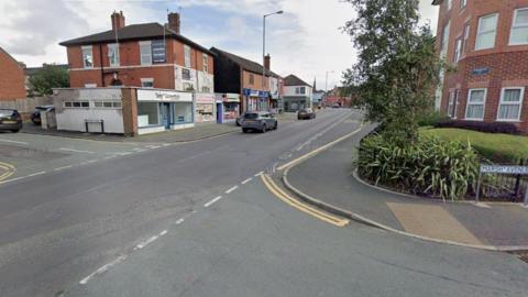 A street view image of a town centre junction. Cars can be seen driving along the road, with shops and business premises on the left. A block of flats is behind a black fence on the right, with a tree near the fence. A street sigh next to the fence says "Marsh Avenue".