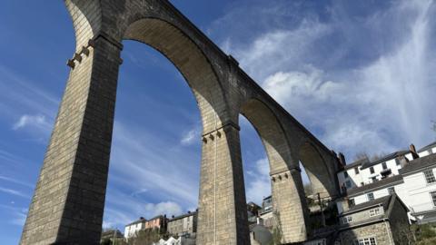 Looking up at the arches of the viaduct from a location on the Tamar River. There are houses nestled into the hillside and around part of the viaduct. There is blue sky 