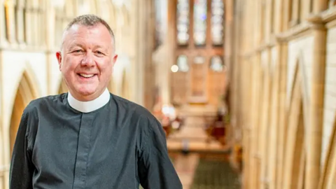 Father Simon Robinson who has short grey hair in his clerical collar standing inside Truro Cathedral with the altar blurred behind him in the background. He is smiling.