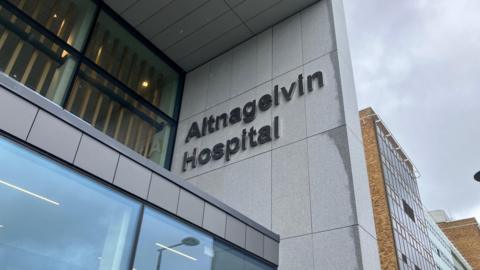 A close up of a sign outside Altnagelvin Hospital, which says the name of the building in large lettering. It is a tall building with a mix of facades including brown brick and grey cladding.