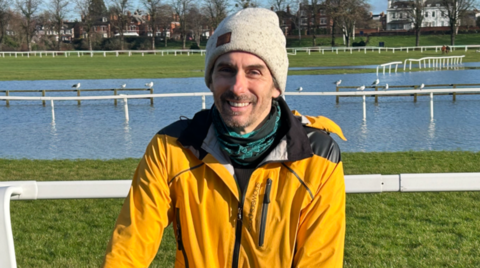 A man wearing a yellow coat is looking at the camera. White railings, water and trees are in the background.